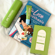 Personal Day skincare and beauty products arranged on a white fabric background, including a green cylindrical bottle, a green tube with a blue cap, a blue book titled 'The Love Boothesis', and a green boxed item labeled 'Personal Day'.