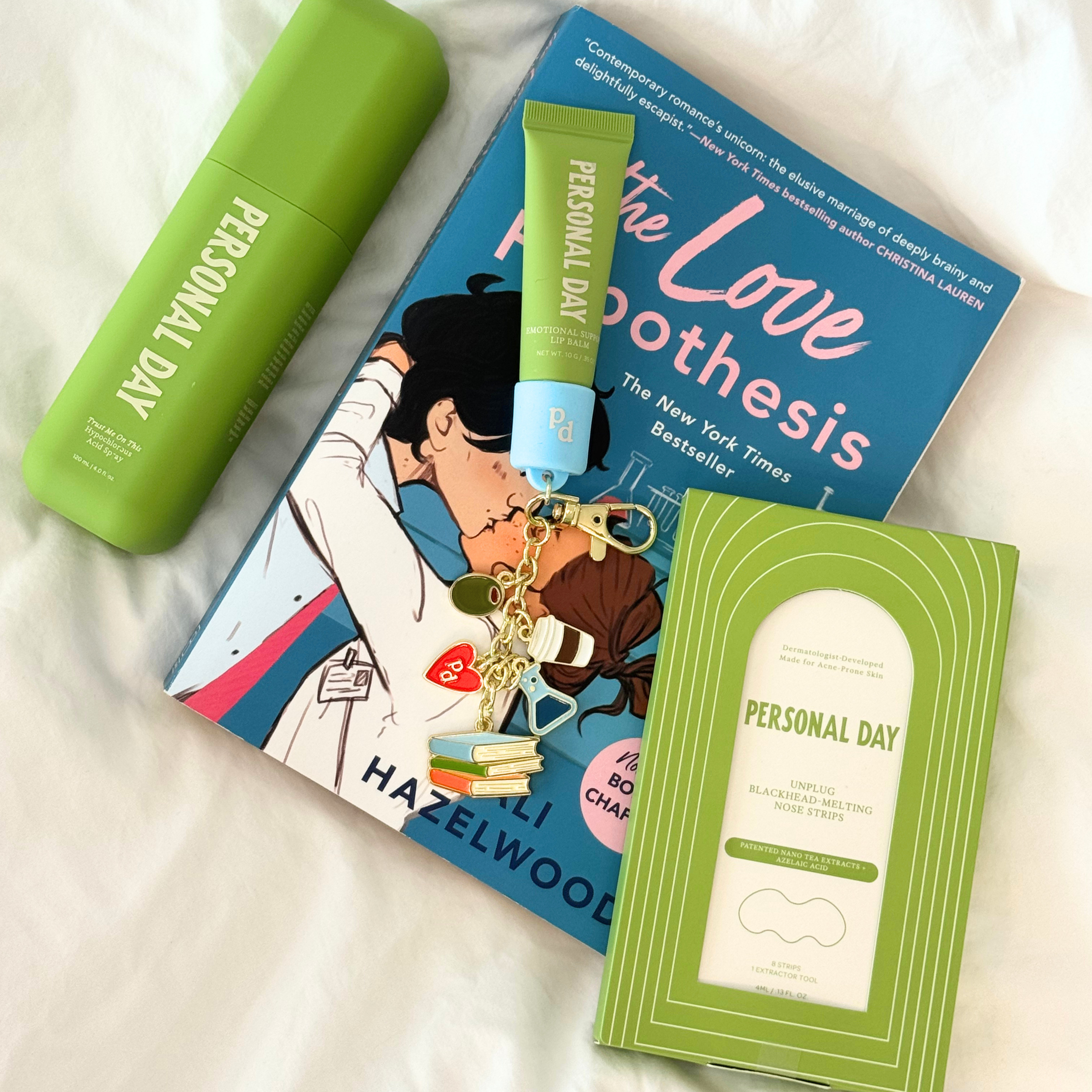 Personal Day skincare and beauty products arranged on a white fabric background, including a green cylindrical bottle, a green tube with a blue cap, a blue book titled 'The Love Boothesis', and a green boxed item labeled 'Personal Day'.