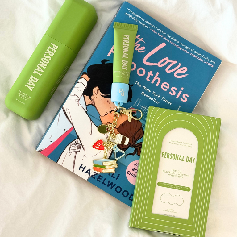 Personal Day skincare and beauty products arranged on a white fabric background, including a green cylindrical bottle, a green tube with a blue cap, a blue book titled 'The Love Boothesis', and a green boxed item labeled 'Personal Day'.